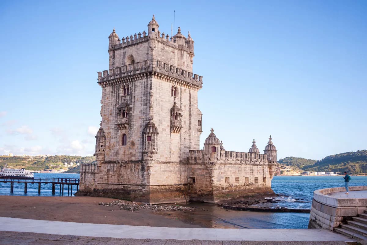 The Tower of Belém in Lisbon, a carved stone tower with turrets, standing at the edge of the River Tagus under a blue sky