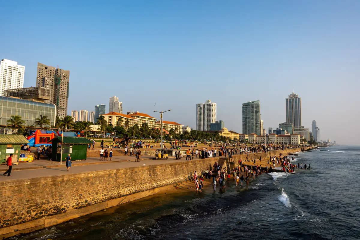 People strolling along the Galle Face seafront promenade in Colombo, with a sea wall, waves and tall buildings behind