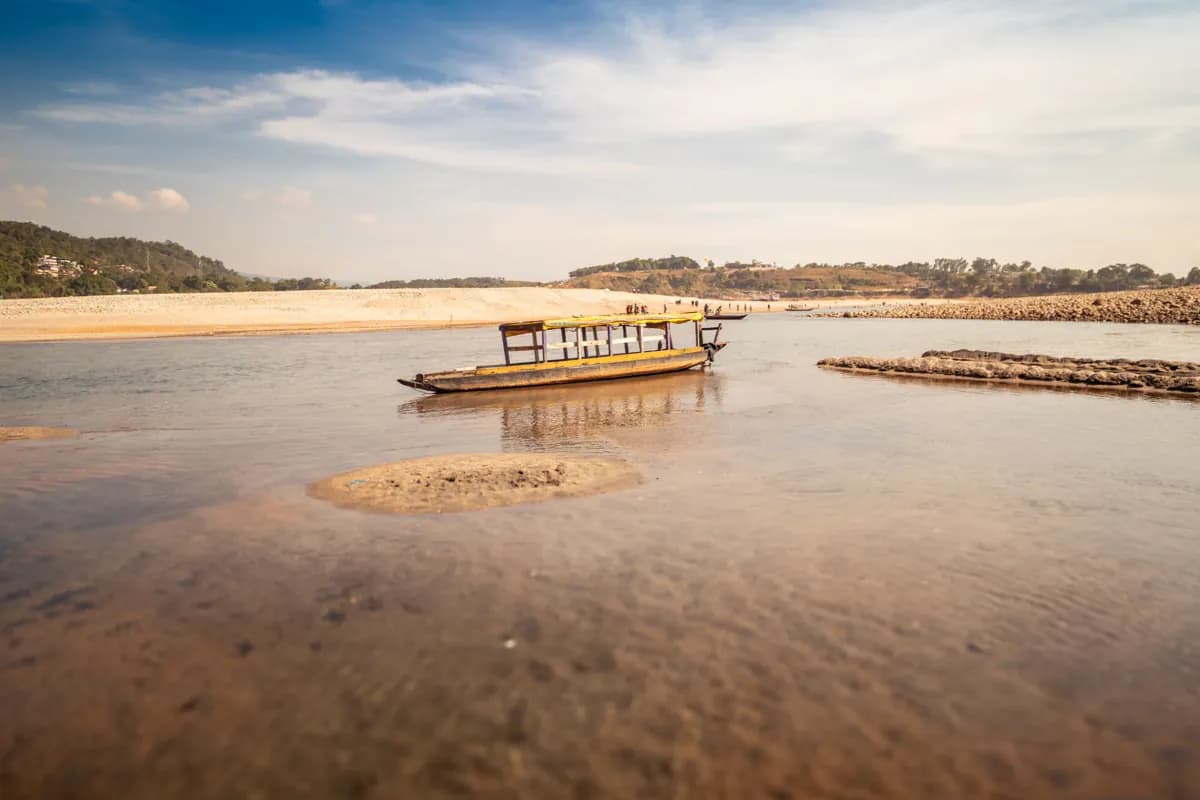 A wooden boat in the shallow river at Jaflong, with stony banks and green hills beyond