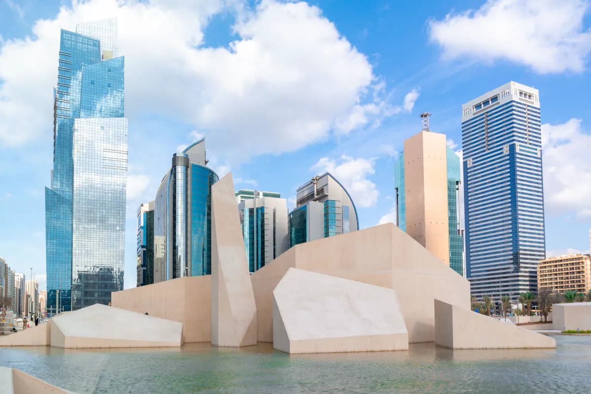 Angular pale stone forms rising from a shallow pool in front of glass office towers in Abu Dhabi