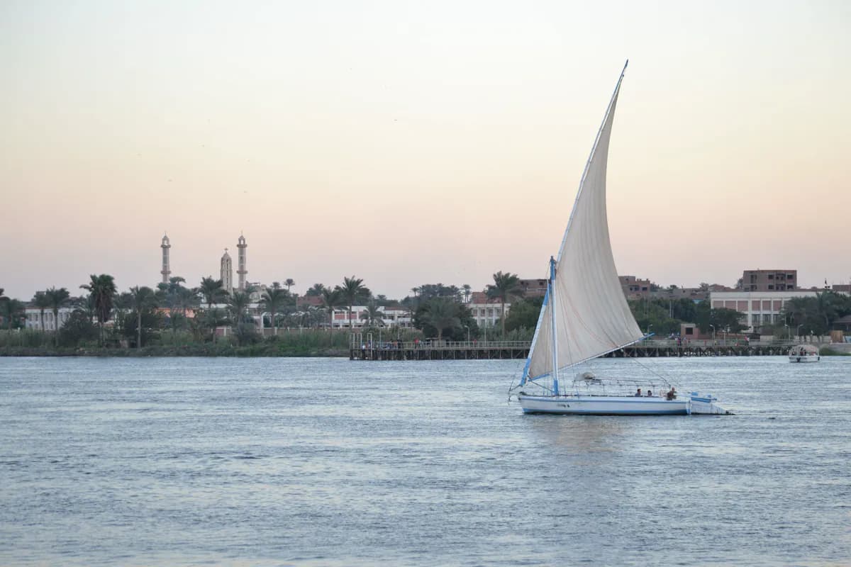A felucca with a white sail on the Nile at dusk, with palm trees, minarets and a church tower on the far bank