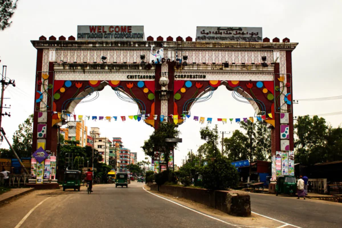 A colourful city entrance arch over a road in Chattogram, with auto rickshaws and a cyclist passing beneath