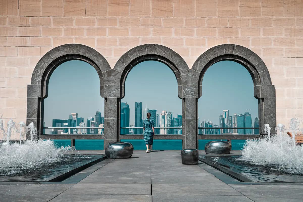 Three stone arches at the Museum of Islamic Art framing the Doha skyline across the water, with fountains on either side