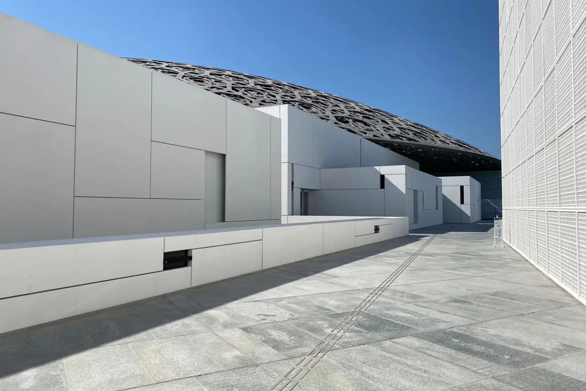 White gallery blocks and a paved terrace beneath the patterned metal dome of Louvre Abu Dhabi under a blue sky
